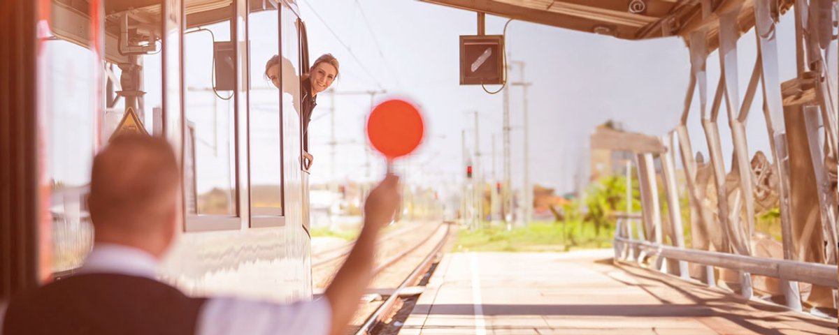 Eine Triebfahrzeugführerin schaut aus dem Zugfensters des Führerstands auf den Kundenbetreuer, der eine rote Signalkelle in die Luft hebt. 